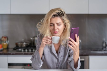 Téléchargez les photos : Triste femme bavardant sur un téléphone portable dans la cuisine à la maison. Femme déprimée dans la cuisine le matin. Ménagère mauvaise humeur le matin - en image libre de droit
