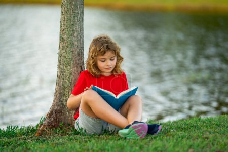 Foto de Lindo niño leyendo libro en el prado al aire libre. Escuela y educación. Naturaleza y parque. Aprendizaje temprano. Un niño leyendo un libro en el parque de verano. Concepto de recreación infantil sin artilugios - Imagen libre de derechos