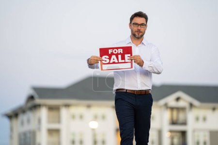 Photo for Successful real estate agent in a suit holding for sale sign near new apartment. Real estate agent with home loan contract, selling home. Realtor or real estate agent shows board for sale - Royalty Free Image