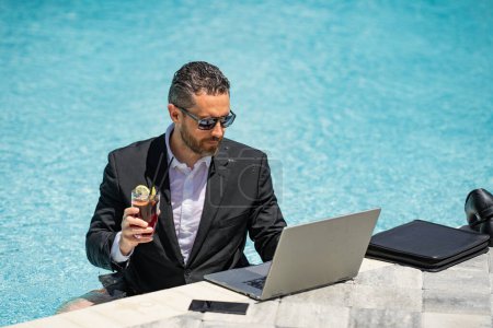 Téléchargez les photos : Les affaires d'été. Homme d'affaires drôle en costume avec ordinateur portable dans l'eau de la piscine. Des affaires d'été à distance. Homme d'affaires relaxant dans la piscine en costume. Travail indépendant, commerce en ligne. Affaires à distance - en image libre de droit