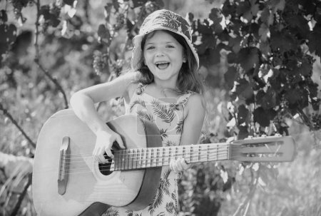 Foto de Linda niña con cabello rubio rizado tocando la guitarra en el fondo de la naturaleza. Cara de niños soñadores. Sonriente niño jugando al aire libre en verano - Imagen libre de derechos
