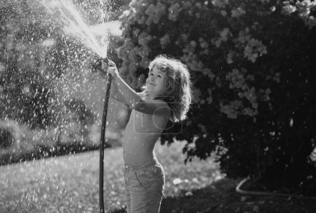 Téléchargez les photos : Enfant drôle amusez-vous et souriant heureux sur la nature arrière-cour. Arrosage des plantes dans le jardin à la maison le jour de l'été - en image libre de droit
