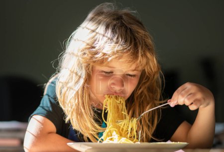 Téléchargez les photos : Gros plan portrait d'un gamin drôle mangeant des nouilles spaghetti aux pâtes. Petit garçon petit déjeuner dans la cuisine - en image libre de droit
