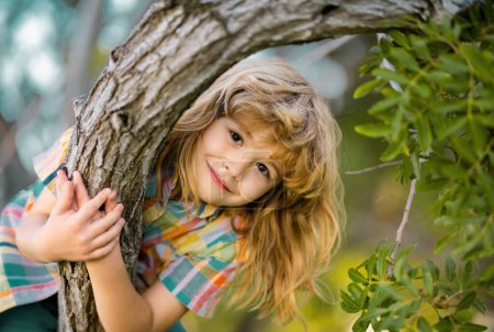 Téléchargez les photos : Mignon petit garçon appréciant grimper sur l'arbre le jour de l'été. Enfant heureux couché dans un arbre embrassant une grande branche - en image libre de droit