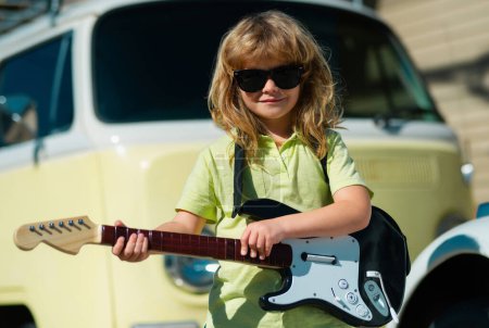 Foto de Niño divertido con pelo rubio rizado tocando la guitarra sobre fondo amarillo beige. Divertido pequeño músico hipster niño tocando la guitarra - Imagen libre de derechos