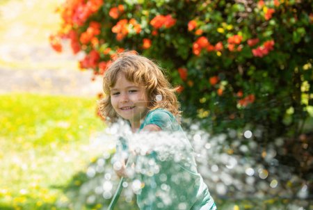 Téléchargez les photos : Le gamin qui joue dans le jardin, verse du tuyau, fait pleuvoir. Joyeux concept d'enfance. Un enfant verse de l'eau sur les arbres. Soin des enfants pour les plantes dans la cour arrière - en image libre de droit