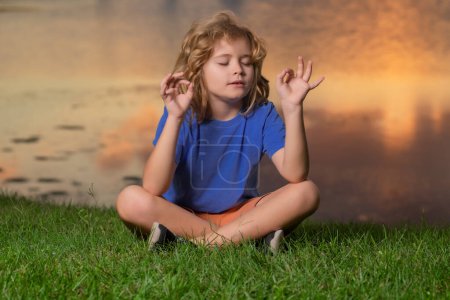 Foto de Niño con la atención cercana a los ojos meditando, sentarse en la hierba para la paz y el yoga. Niño relajarse en la puesta de sol de verano. Concepto de libertad y niños despreocupados - Imagen libre de derechos