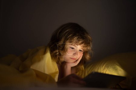Téléchargez les photos : Enfant jouant jeu sur tablette au lit la nuit. Des enfants avec des réseaux sociaux. Enfant couché au lit jouant une tablette dans une pièce sombre, lumière sous le noir. Gros plan de l'enfant regarder des dessins animés sur tablette - en image libre de droit