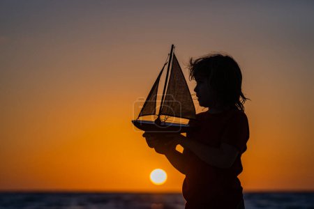 Photo for Silhouette of kid playing with toy seailing boat on sunset sea. Little kid boy sailing toy ship on sea water. Summer vacation with kids. Kid dreaming about sailing - Royalty Free Image