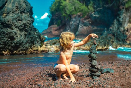 Téléchargez les photos : Le gamin construit un cairn de pierres sur la côte. Jeux de plage avec les enfants. Loisirs en soirée en vacances avec les enfants. Équilibre développement bâtiment fantaisie. Enfance. Chalut pour enfants de l'océan - en image libre de droit