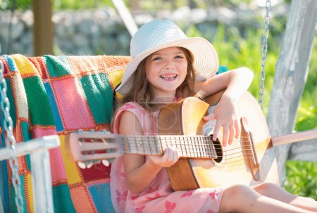Téléchargez les photos : Enfant musicien jouant de la guitare comme une rockstar. Activité estivale pour les enfants par temps chaud. Petite fille mignonne s'amuser sur une balançoire dans un beau jardin d'été et journée ensoleillée en plein air - en image libre de droit