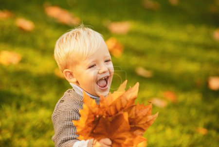 Foto de Niño emocionado jugando con hojas en el parque de otoño. Niño rubio sonriente sostiene hojas de otoño en la naturaleza. Sonríe feliz cara de niños. Concepto de infancia feliz - Imagen libre de derechos