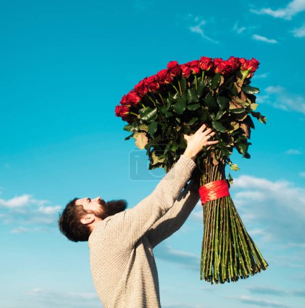 Foto de Hombre con flores de ramo de rosas. El hombre barbudo tiene un gran ramo de rosas rojas, proponerle matrimonio a su novio en la cita. Romántico chico celebrar el día de San Valentín - Imagen libre de derechos