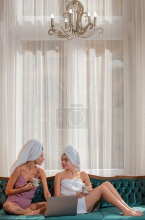 Foto de Happy young women wearing bathrobes and towel on heads celebrating wedding or birthday party, sitting on bed - Imagen libre de derechos