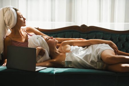 Téléchargez les photos : Deux jeunes belles femmes en peignoirs blancs et serviettes sur la tête au lit après le réveil. Modèles sexy insouciants sur le lit dans l'appartement ou la chambre d'hôtel. Repos matinal, calme-toi - en image libre de droit