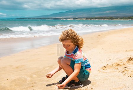 Foto de Lindo chico jugando con arena en la playa tropical de verano. Niño feliz sentado en la playa de arena y jugando con la arena - Imagen libre de derechos