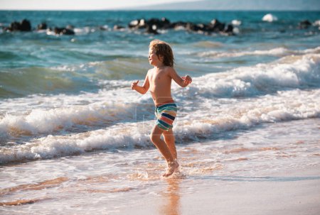 Téléchargez les photos : Petit garçon courant sur le rivage de la plage éclaboussant l'eau dans la mer bleue. Enfant marchant sur la plage d'été - en image libre de droit
