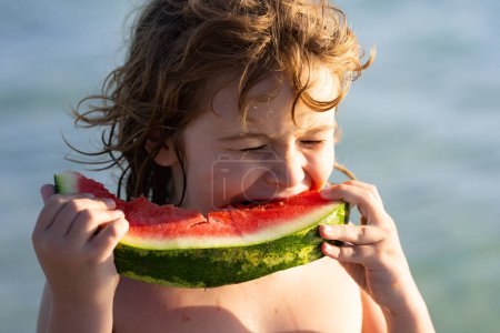 Téléchargez les photos : Le petit enfant mange de la pastèque rouge juteuse sur la plage. Vacances d'été pour enfants, fruits de saison et baies. Portrait extérieur d'enfant d'été. Face d'enfant d'été. Enfant à la pastèque, fruits d'été - en image libre de droit