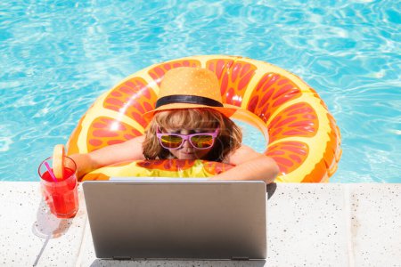 Téléchargez les photos : Boire un cocktail d'été pour enfants et utiliser un ordinateur portable dans la piscine. Enfant travaillant sur ordinateur portable dans la piscine. Tourisme de voyage et concept d'affaires. Blogues d'été - en image libre de droit