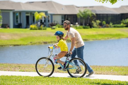 Photo for Father teaching son riding bike. Father helping excited son to ride a bicycle in american neighborhood. Child in bike helmet learning to ride cycle with his dad. Fathers day - Royalty Free Image