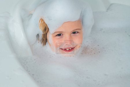 Foto de Cara de niños en espuma. Niño divirtiéndose en el baño con burbujas. Niño feliz disfrutando del baño. Niño sonriendo en el baño con espuma de jabón. El niño se baña en un baño con espuma - Imagen libre de derechos