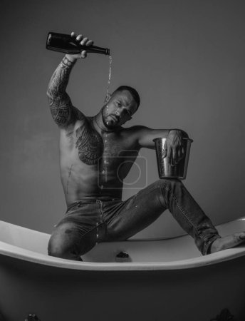 Téléchargez les photos : Homme tatoué musclé fort tenant bouteille de champagne et posant dans la salle de bain. Beau barbu homme torse nu en jeans avec corps sexy dans la salle de bain. macho homme sexuel dans le bain - en image libre de droit