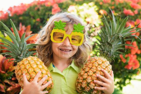 Foto de Niño con gafas de sol divertidas con piña. Niño con fruta tropical. Vacaciones exóticas. Fruta para niños. Vacaciones de verano para niños - Imagen libre de derechos