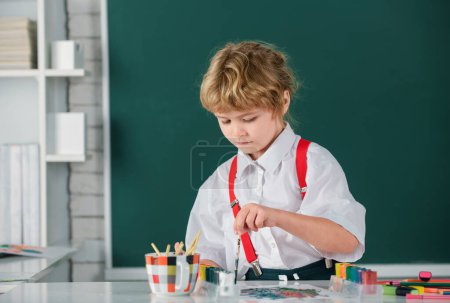 Foto de El niño pequeño dibuja en el aula sentado en una mesa, divirtiéndose en el fondo de la pizarra escolar. Educación de los niños sobre artes y oficios - Imagen libre de derechos