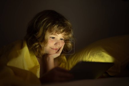 Téléchargez les photos : Le gamin est accro à la tablette, le gamin utilise la tablette, regarde le dessin animé. Enfant regardant sa tablette dans le lit. Visage enfant éclairé à partir de l'écran de l'appareil. Garçon sous les couvertures tenir une tablette. Temps de nuit - en image libre de droit