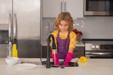 Téléchargez les photos : Enfant garçon laver la vaisselle dans l'évier de la cuisine. Détergents et accessoires de nettoyage. Service de nettoyage. petit garçon ménage - en image libre de droit