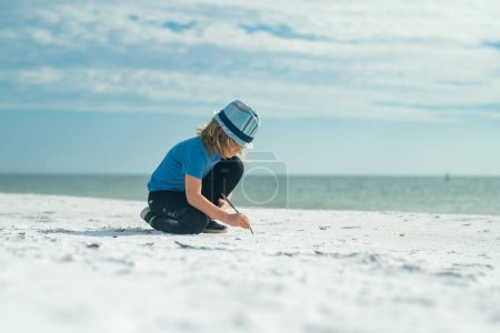 Téléchargez les photos : Joyeux enfant dessinant sur un sable dans la mer d'été. Un gamin qui s'amuse à la plage. Vacances d'été et enfants actifs. Voyage et concept d'aventure estivale. Enfant écrire dans le sable en vacances d'été - en image libre de droit
