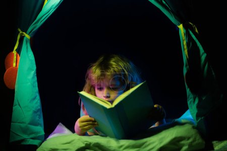 Foto de Niños en tienda de campaña niños leyendo un libro en la casa oscura. Niño leyendo un libro acostado en la cama. Niño soñando leer cuentos, cuentos de hadas o cuentos de hadas - Imagen libre de derechos