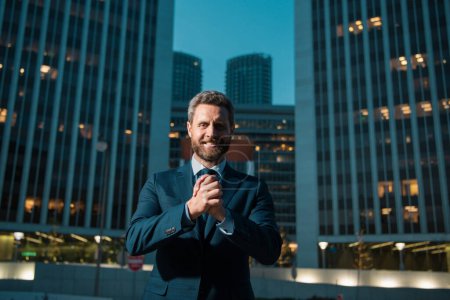 Téléchargez les photos : Heureux homme d'affaires souriant en costume près du bureau, employeur réussi à faire un marché tout en se tenant près du bureau gratte-ciel la nuit - en image libre de droit