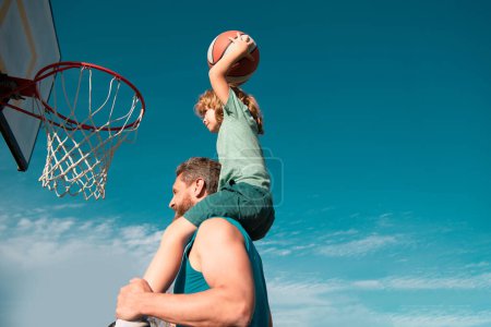 Photo for Father and son playing basketball. Sports Concept. Boy child sitting on the dad shoulders, throwing basketball ball into basket, side view on sky background with copy space - Royalty Free Image