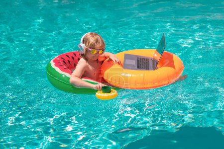 Téléchargez les photos : Enfant travaillant sur ordinateur portable à la piscine. Technologie en ligne estivale. Voyageur relaxant sur la plage de la mer tropicale pendant les vacances d'été. Voyage, enfant utilisant un ordinateur portable - en image libre de droit