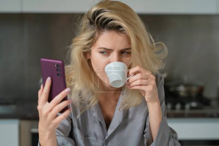 Téléchargez les photos : Fatiguée femme malheureuse bavarder sur téléphone portable sur la cuisine à la maison. Humeur de femme au foyer le matin - en image libre de droit