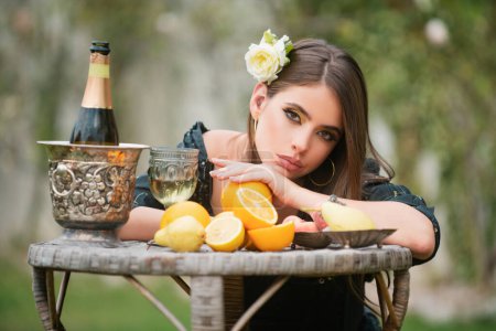 Foto de Retrato de chica sexy en el picnic de verano en una hierba en el parque con vino y frutas frescas. Foto de moda al aire libre de la hermosa mujer joven con rosa en el pelo. Belleza mujer cara - Imagen libre de derechos