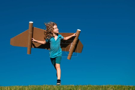 Foto de Pequeño niño juega astronauta o piloto. Niño corriendo sobre el fondo del cielo azul. Niños con alas de papel jetpack sueños. Los niños imaginan sueños de volar. Divertido niño con juguete jet pack - Imagen libre de derechos
