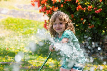 Téléchargez les photos : Arrosage des fleurs et des plantes dans le jardin. Enfant avec tuyau d'eau dans la cour. Jardinage des enfants. Enfants plaisir d'été en plein air à la maison - en image libre de droit