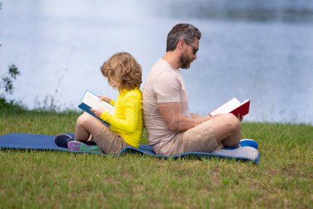 Foto de Padre e hijo disfrutan de pasar tiempo de calidad leyendo libros al aire libre. Papá y su hijo disfrutan de la naturaleza mientras leen libros al aire libre. Padre y su pequeño hijo leyendo un libro en Park. Día de los padres - Imagen libre de derechos