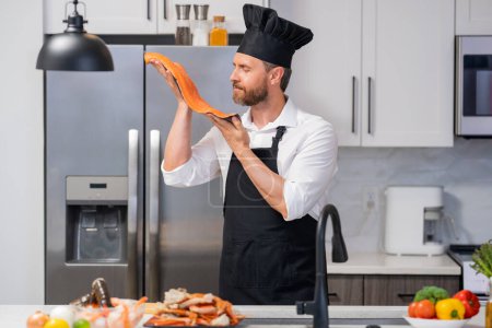 Photo for Handsome senior man in cook apron and chef hat cooking fish salmon seafood in kitchen. Portrait of middle aged man cooking seafood in kitchen. Millennial man preparing raw fish salmon and seafood - Royalty Free Image
