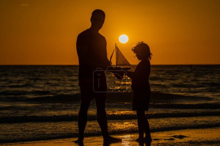 Foto de Padre e hijo jugando al atardecer en el mar. Padre e hijo juegan en el mar con barco de juguete al amanecer. Silueta de padre e hijo. Padre e hijo divirtiéndose en la playa del atardecer. Día de los padres - Imagen libre de derechos