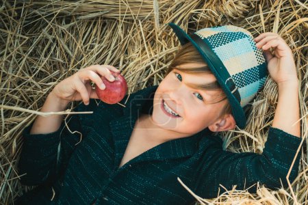 Téléchargez les photos : Le garçon s'allonge sur un fond de foin et mange une pomme. Rêve d'automne. Rêves d'enfants sur la nature d'automne. Concept de rêve d'enfance. Enfant rêveur. Rêves et imagination. Visage d'enfant rêveur - en image libre de droit