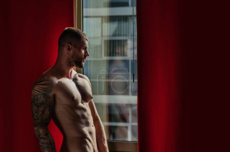 Muscular male torso, bare shoulders. Nude man in a bedroom. Young sexy body of strong man at morning. Muscular man in hotel room on window curtains. Shirtless topless sexy male model posing indoor