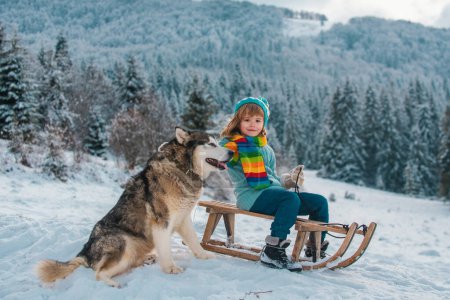 Téléchargez les photos : Garçon drôle avec chien s'amuser avec un traîneau en hiver. Enfants avec chien loup. Concept de vacances d'hiver. Amour animal de compagnie - en image libre de droit