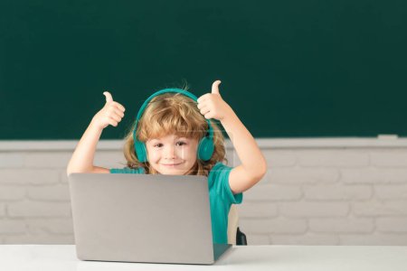 Photo for Clever school boy with thumbs up, cute pupil wears headphones writing on laptop, listen audio lesson use computer. Videocall, e-study with tutor distantly. First day at school - Royalty Free Image
