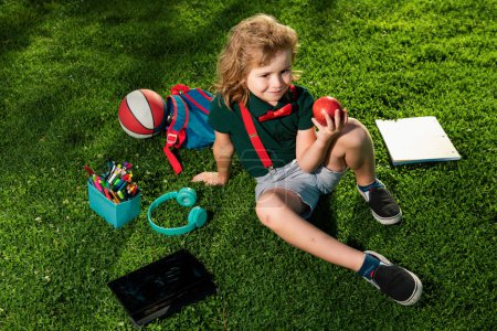 Foto de Niño con útiles escolares sentado al aire libre en el jardín, sonriendo alegremente disfrutando del tiempo libre, vistiendo pantalones cortos y camisa - Imagen libre de derechos