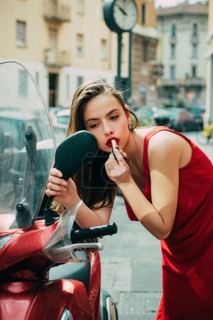 Téléchargez les photos : Portrait de belle mannequin sexy fille de mode avec lèvres rouges mettre rouge à lèvres regardant dans un miroir voiture de conduite. Rouge à lèvres rouge blonde regardant dans le miroir sur un cyclomoteur. visage d'une belle femme brune dans - en image libre de droit