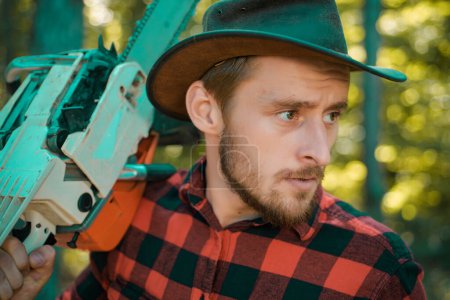 Foto de Lumberjack - retrato de cerca. Un leñador con motosierra en las manos. Cortador de madera con hacha o motosierra en el bosque de verano. Trabajador de madera caminando en el bosque con motosierra - Imagen libre de derechos