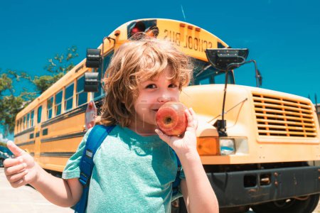 Foto de Niño de la escuela primaria con bolsa en el autobús escolar backgroung. Niño de la escuela en la parte delantera del autobús escolar con manzana - Imagen libre de derechos